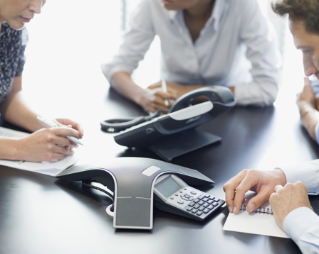Three business professionals are seated around a conference table, writing notes while participating in a meeting.